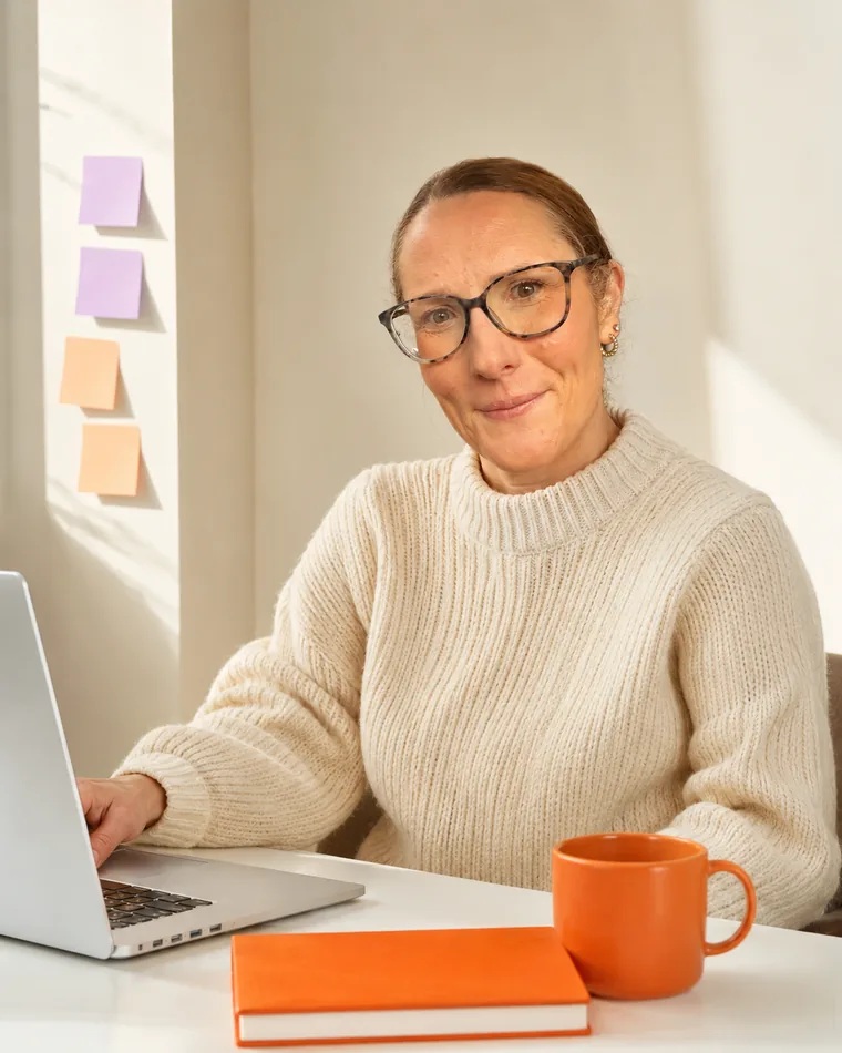 Rachel at her desk with her laptop open, smiling — an orange notebook and mug beside her, sticky notes on the wall